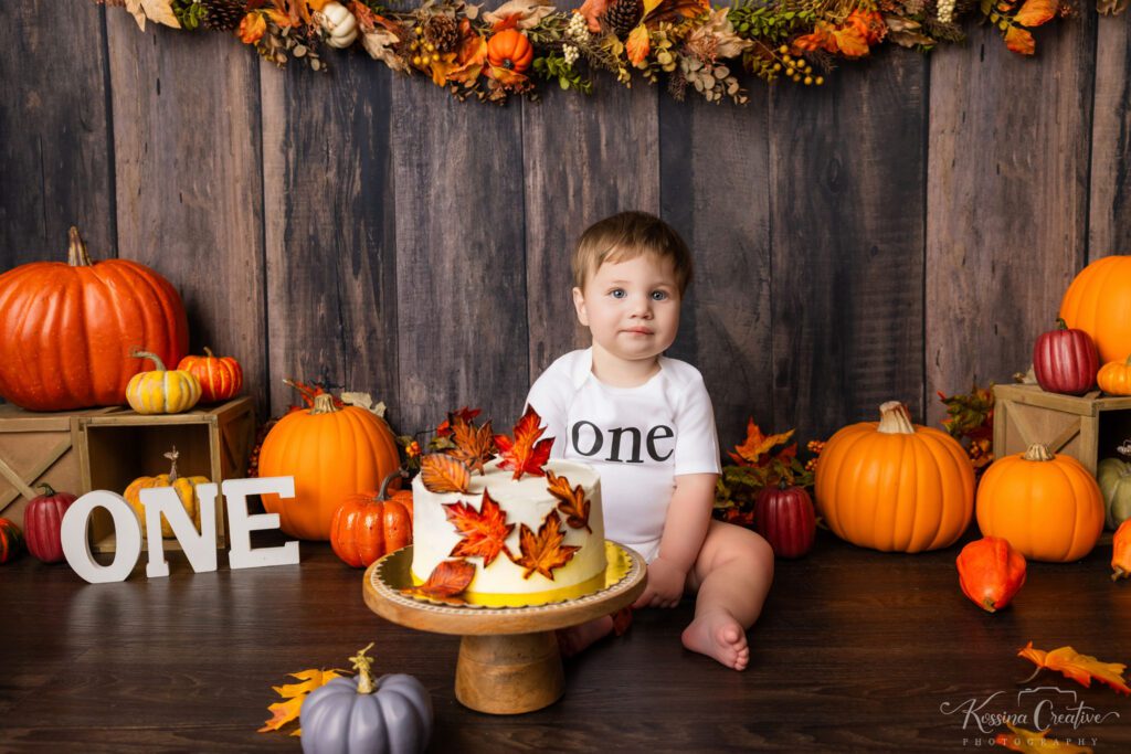Photo of one year old boy with white frosted cake, dark wood backdrop, and pumpkin decorations in various sizes and shades of orange